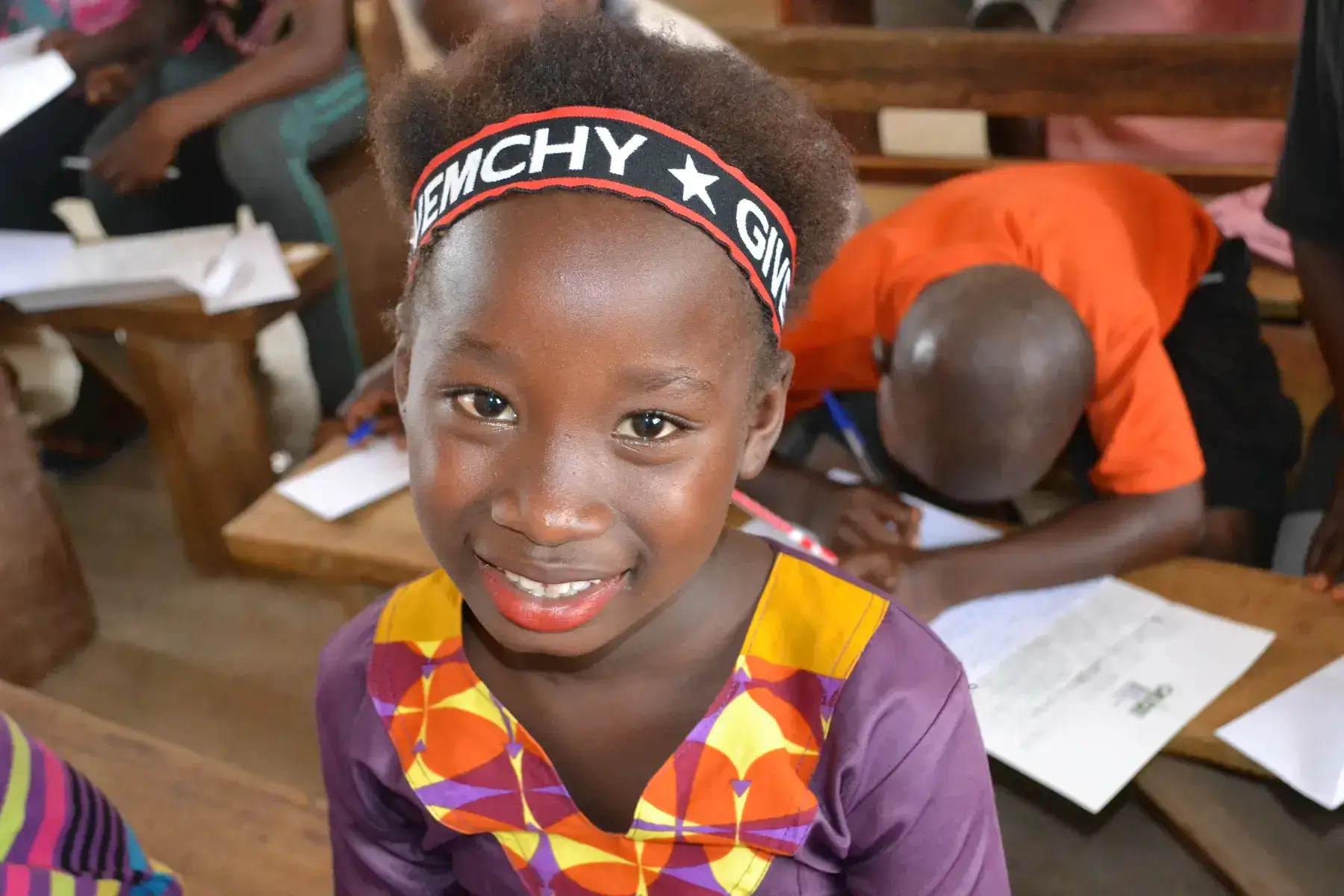 Girl in a classroom, smiling at camera with partial view of other students in background.